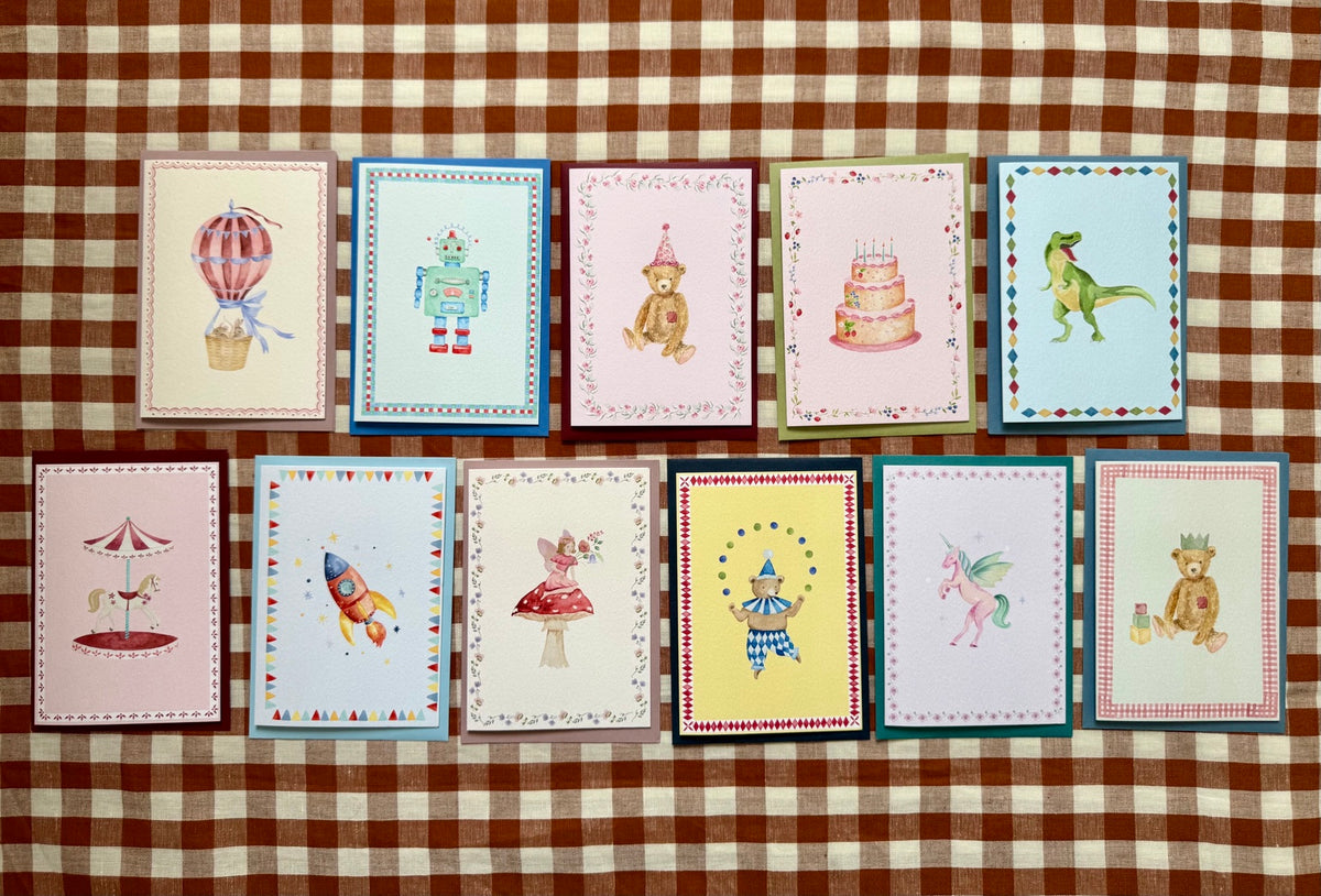 Eleven Kate MacLeod Designs hand-painted children's greetings cards arranged in two rows on brown gingham cloth